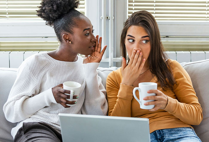 Two women sharing a disturbing secret while holding coffee mugs, sitting closely on a couch with a laptop nearby.