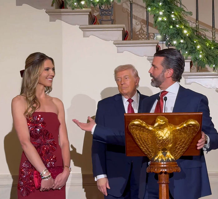 Donald Trump Jr. speaking at podium during engagement event with woman in red dress and man in dark suit.