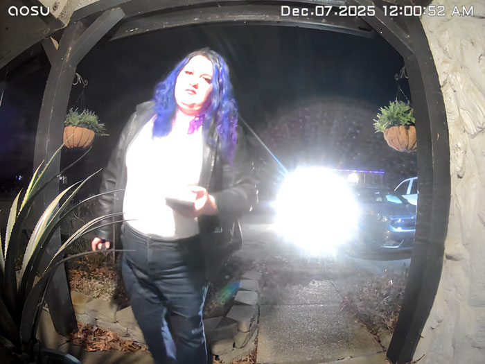 Person with blue hair standing at front door captured by porch camera with bright headlights in background at night.
