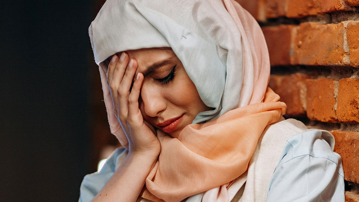 Young Muslim woman wearing hijab, upset and covering face with hand, emotional moment indoors by brick wall.
