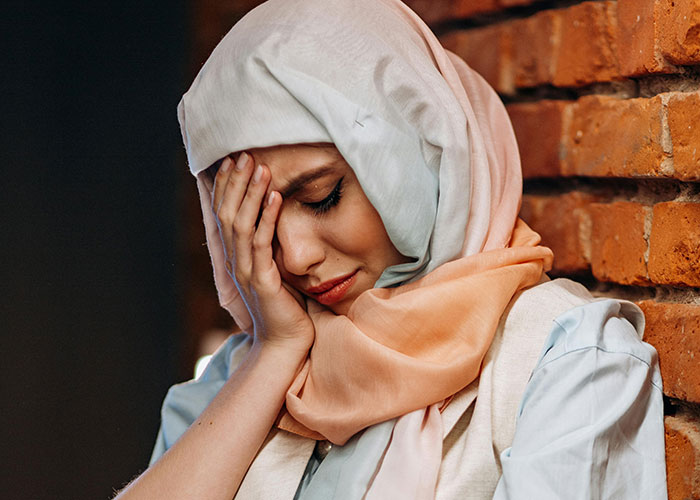 Young Muslim woman wearing hijab, looking distressed and leaning against a brick wall, highlighting family conflict over bridal party attire. Young Muslim woman wearing hijab, looking distressed and leaning against a brick wall, highlighting family conflict over bridal party attire.