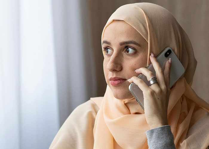 Young Muslim woman wearing a hijab, holding a smartphone, looking concerned while having a phone conversation at home. Young Muslim woman wearing a hijab, holding a smartphone, looking concerned while having a phone conversation at home.
