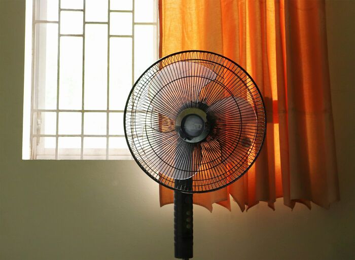 Standing fan in a room near window and orange curtains symbolizing realities of being a postal worker in hot conditions.