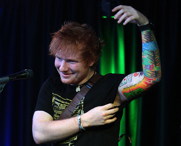 Ed Sheeran smiling on stage, lifting his sleeve to reveal colorful arm tattoos and showing off his physique.