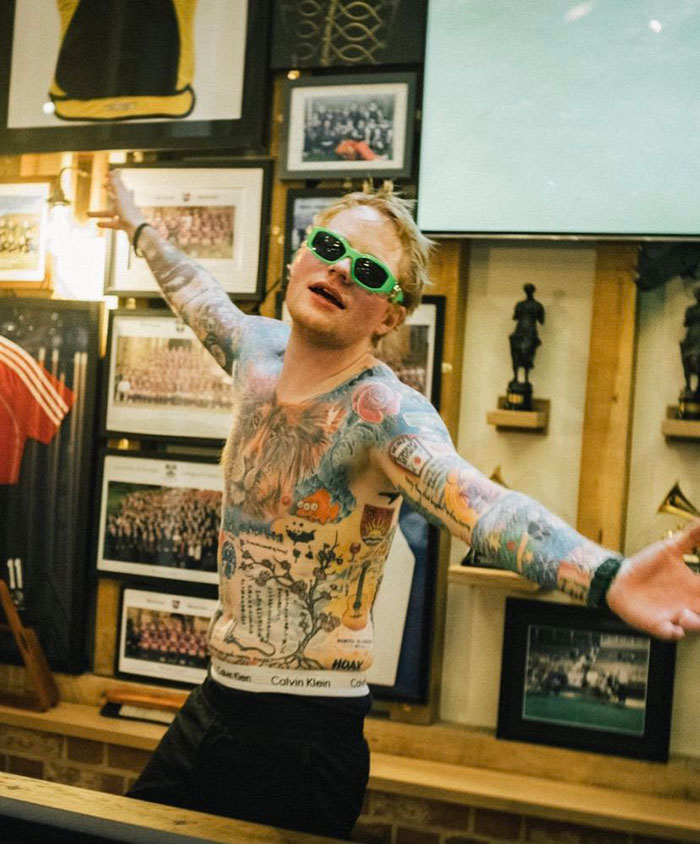 Ed Sheeran with a shredded physique wearing green sunglasses and displaying full-body tattoos indoors.