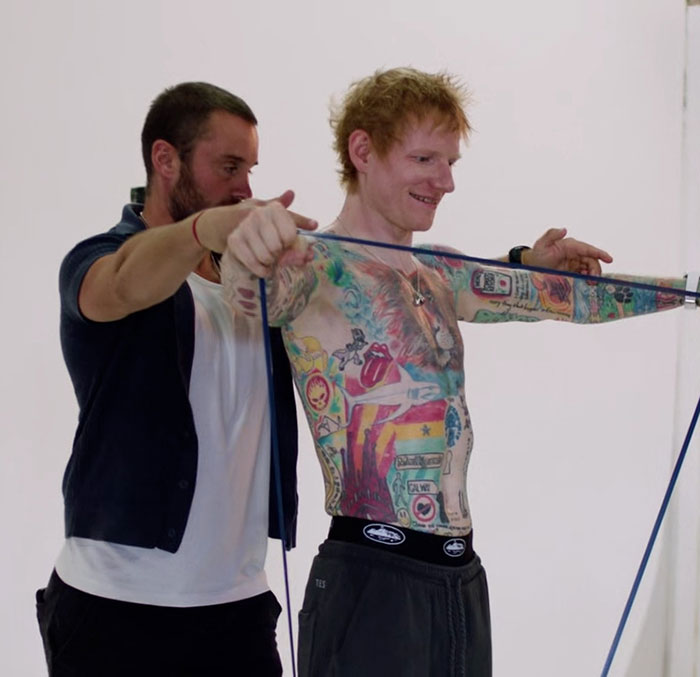 Ed Sheeran shirtless with tattoos, stretching resistance band during fitness training for shredded physique debate