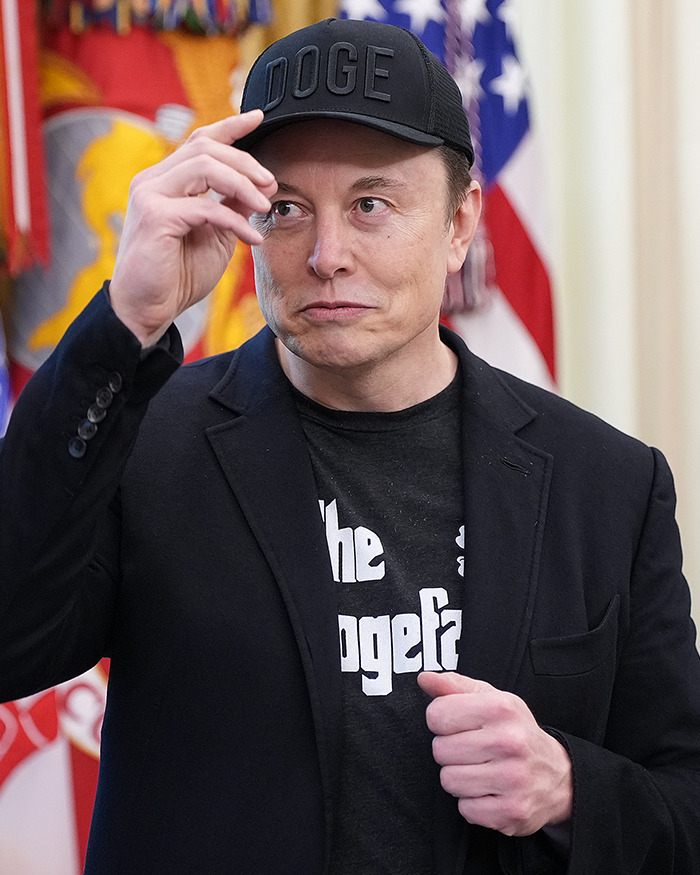 Elon Musk wearing a black Doge hat and jacket, making a gesture with his hand in front of flags indoors. Elon Musk wearing a black Doge hat and jacket, making a gesture with his hand in front of flags indoors.