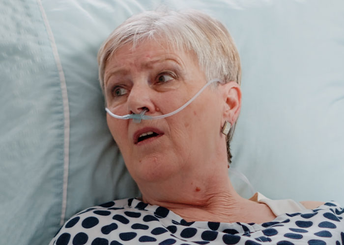 Elderly woman with nasal oxygen tube lying in bed, reflecting on experiences as former cult member.