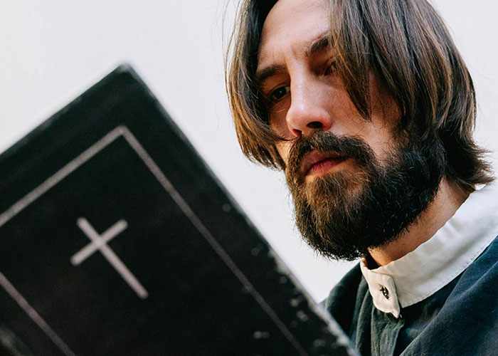 Man with beard reading a black book with a cross, reflecting on the moment they realized they are actually in a cult.