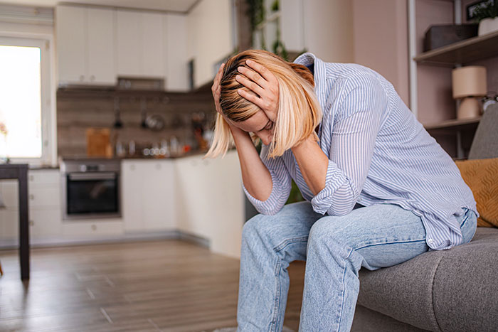 Woman feeling distressed and regretful after Thanksgiving dinner, sitting on a couch with head in hands in a kitchen.