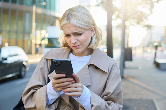 Woman looking upset while checking her phone outdoors, reflecting mom furious after dad and stepmom plan Disney trip excluding her. Woman looking upset while checking her phone outdoors, reflecting mom furious after dad and stepmom plan Disney trip excluding her.