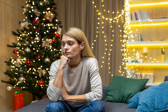 Woman with thoughtful expression sitting on couch near decorated Christmas tree, feeling tired of hosting ungrateful relatives.