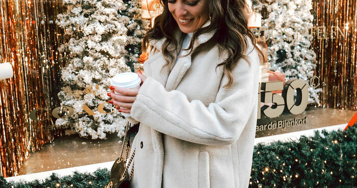 Woman in a white coat holding a cup at a festive European Christmas market with decorated trees and lights in the background