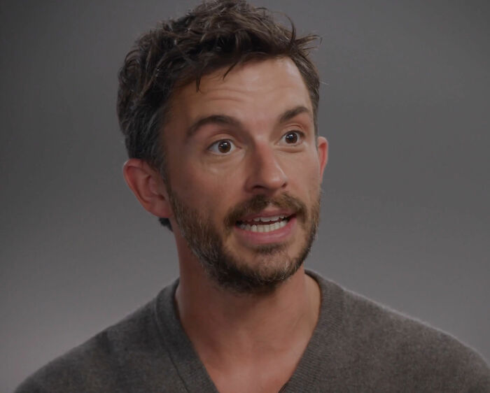 Man with short dark hair and beard speaking, wearing a gray sweater against a plain gray background, discussing masculinity.