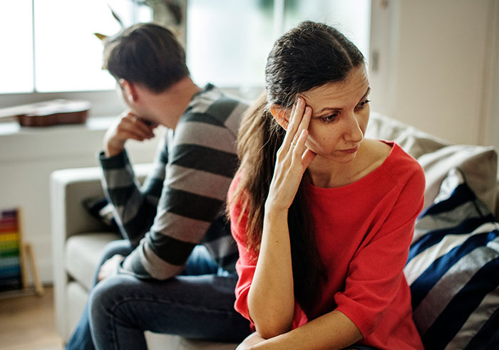 Woman looking distressed at home while man sits turned away, representing restraining order against MIL after long marriage.
