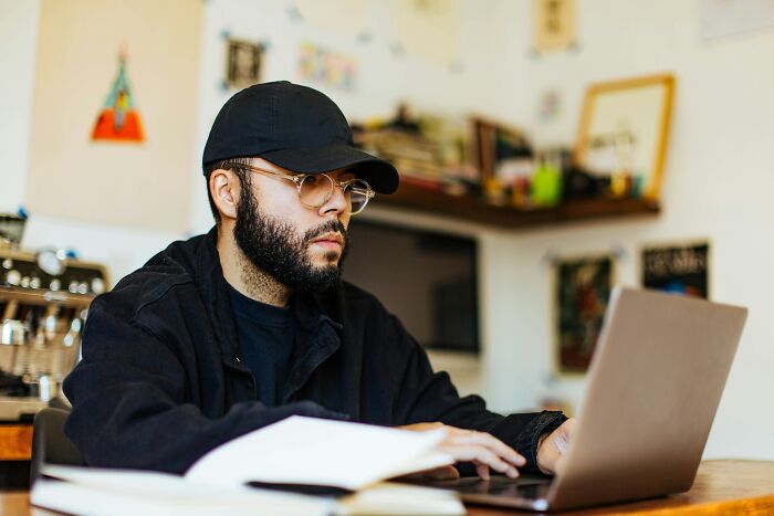 Man with glasses and a cap working on a laptop at home, representing people who became rich out of nowhere.