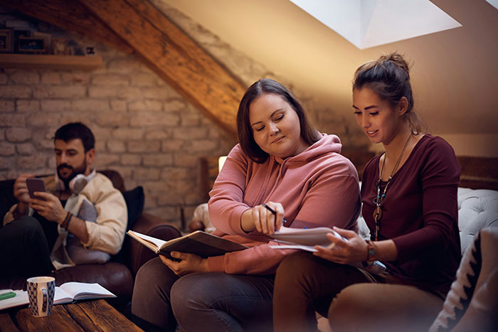 Two friends sitting together indoors, one girl playfully roasting another while the other listens and smiles. Two friends sitting together indoors, one girl playfully roasting another while the other listens and smiles.