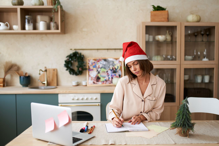 Woman in Santa hat writing in notebook at kitchen table, symbolizing host telling friend's wife about party invite rules. Woman in Santa hat writing in notebook at kitchen table, symbolizing host telling friend's wife about party invite rules.