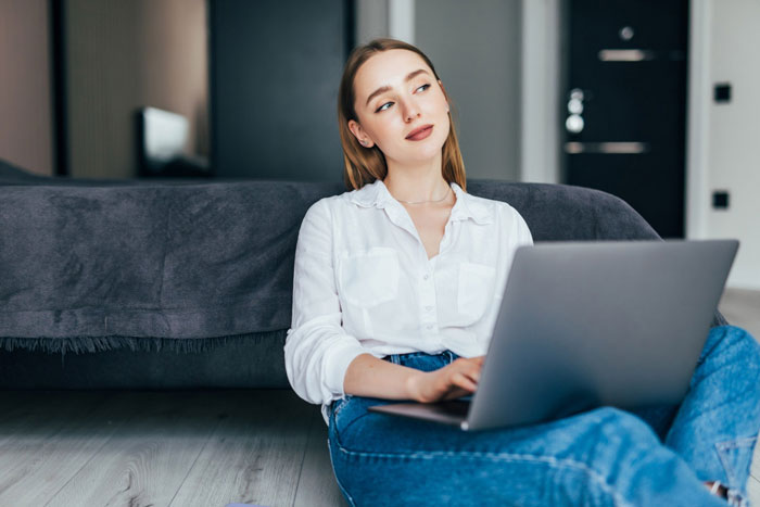 Young woman sitting on floor with laptop looking away, reflecting on host telling friend’s wife about inviting random people. Young woman sitting on floor with laptop looking away, reflecting on host telling friend’s wife about inviting random people.