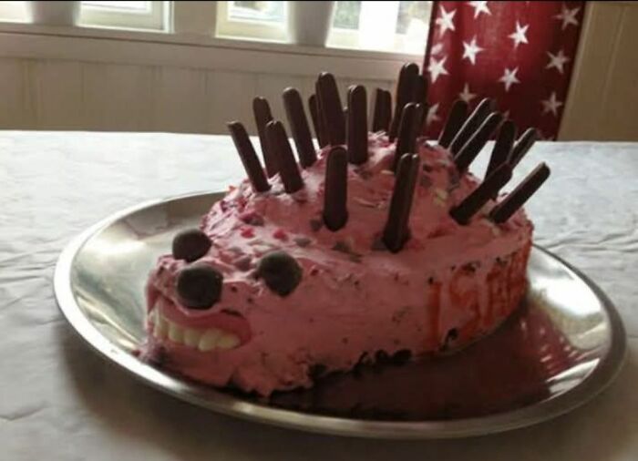 Pink hedgehog-shaped cake with chocolate stick spikes and candy face on a metal tray, featured in food pics to be shamed.