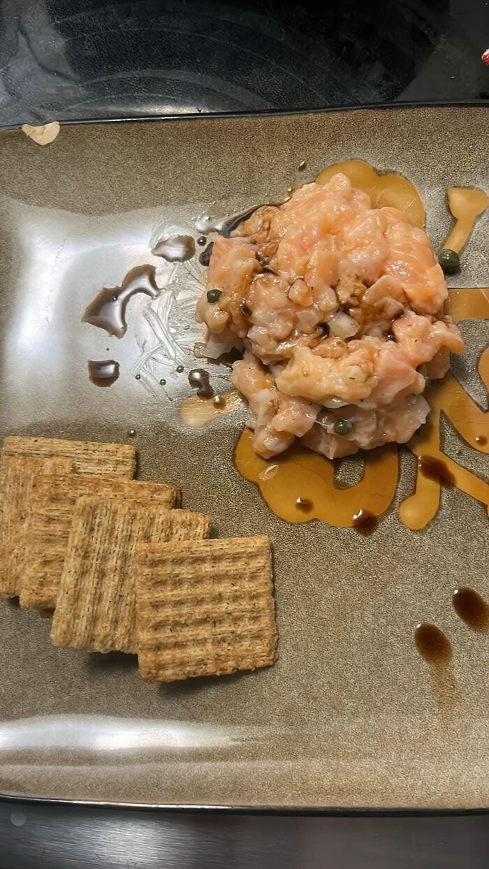 Salmon tartare served with crackers and drizzled sauce on a plate, one of the food pics that deserve to be shamed online.