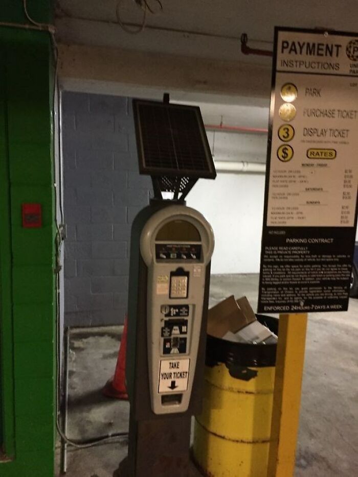 Solar-powered parking meter in a dim garage next to a sign and a yellow trash can, a pointless thing serving no purpose.