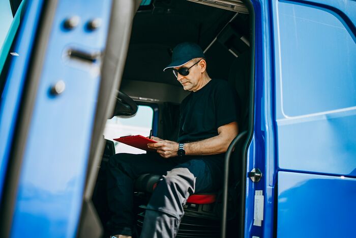 Postal worker in a blue delivery vehicle wearing sunglasses and black clothes, reviewing a clipboard inside the truck cabin.