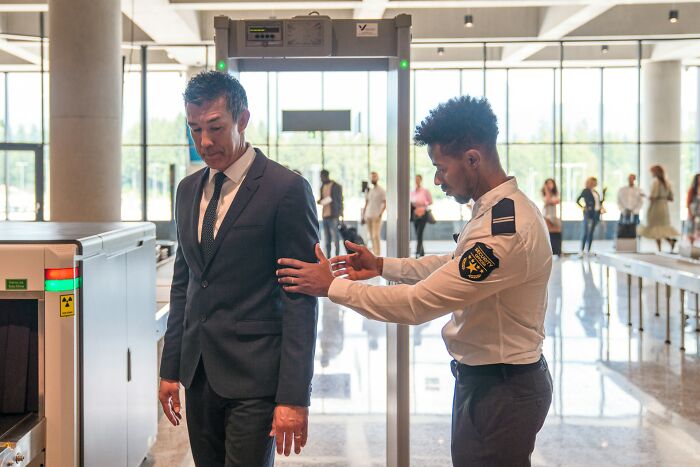 Airport security officer conducting a body check on a passenger near a metal detector in a busy airport terminal.