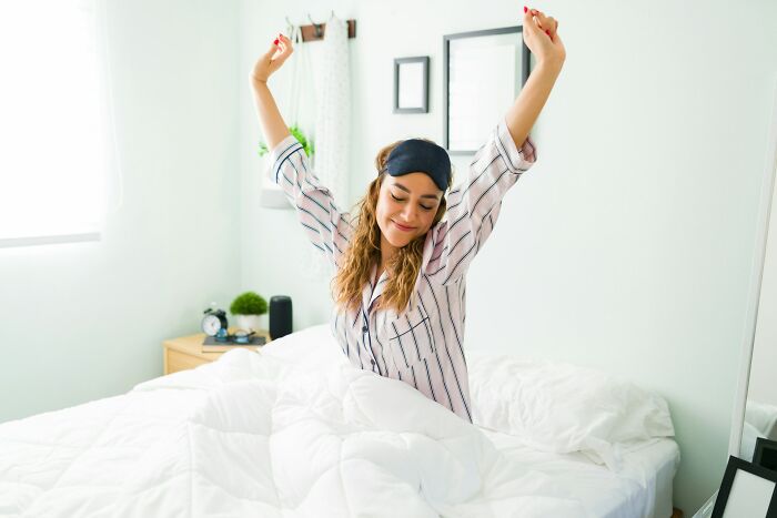 Young woman stretching in bed wearing pajamas and sleep mask, highlighting everyday things more dangerous for health than realized.