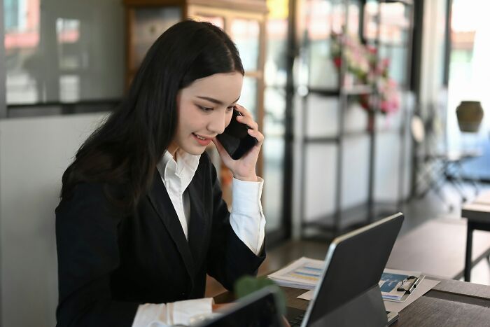 Young businesswoman in formal attire talking on phone while working on laptop in modern office, depicting workplace scandals.