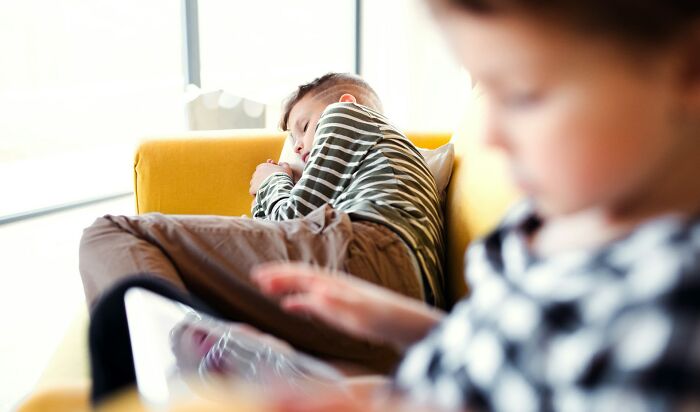 Two children on a yellow couch, one sleeping and the other using a tablet, illustrating modern parenting trends at home.