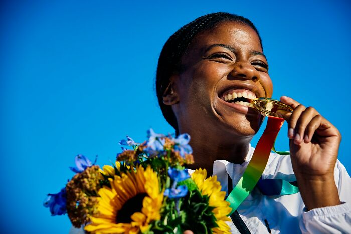 Happy young woman biting gold medal and holding sunflower bouquet, symbolizing victory and success beyond high school era.