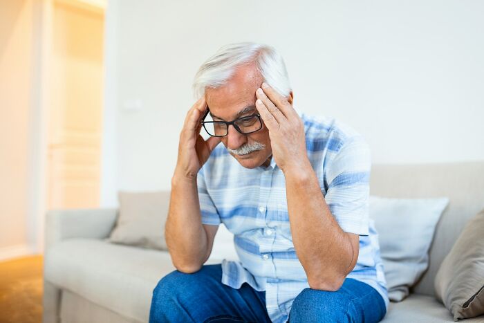 Elderly man sitting on sofa holding head, appearing stressed or thoughtful, illustrating surprising life facts.