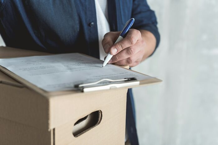 Postal worker filling out forms on a clipboard placed on a cardboard box in a work environment.