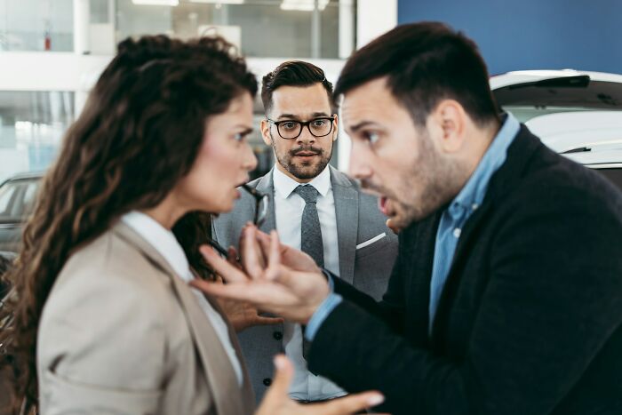 Three professionals in a workplace dispute, with two arguing intensely and a third person observing, depicting workplace scandals.