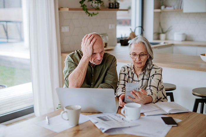Elderly couple reviewing bills at a kitchen table with a laptop, showing frustration and concern about finances. Elderly couple reviewing bills at a kitchen table with a laptop, showing frustration and concern about finances.