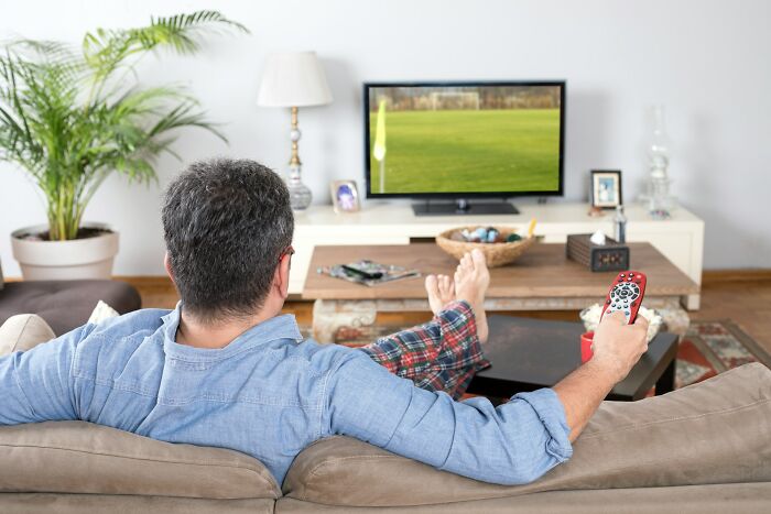 Man sitting on couch watching TV holding remote control, illustrating everyday things more dangerous for health risks.