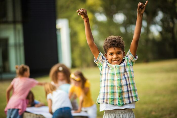 Happy young boy raising hands outdoors while other children play in the background, illustrating dark secrets discovery.