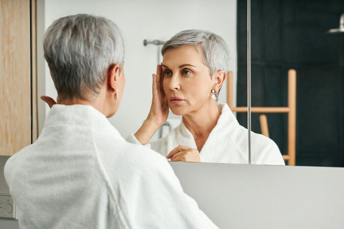 Mature woman in white robe examining her face in a mirror, reflecting on mysteries and unanswered questions.