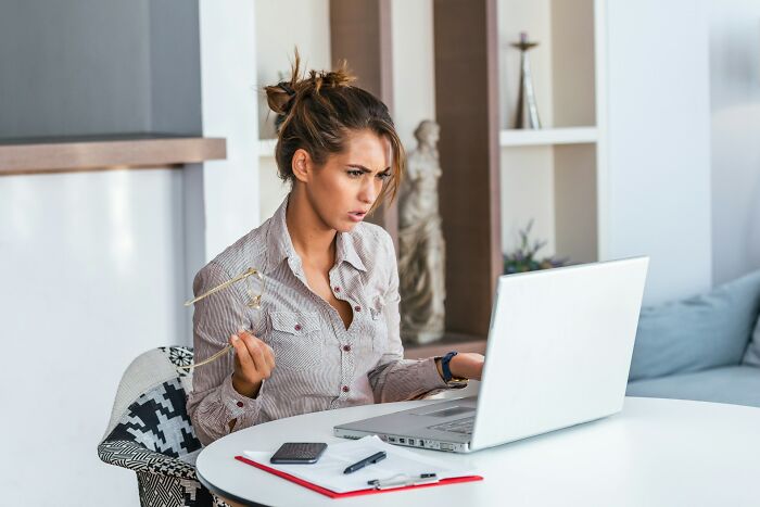 Woman in casual shirt holding glasses, looking shocked while working on a laptop about workplace scandals and issues.