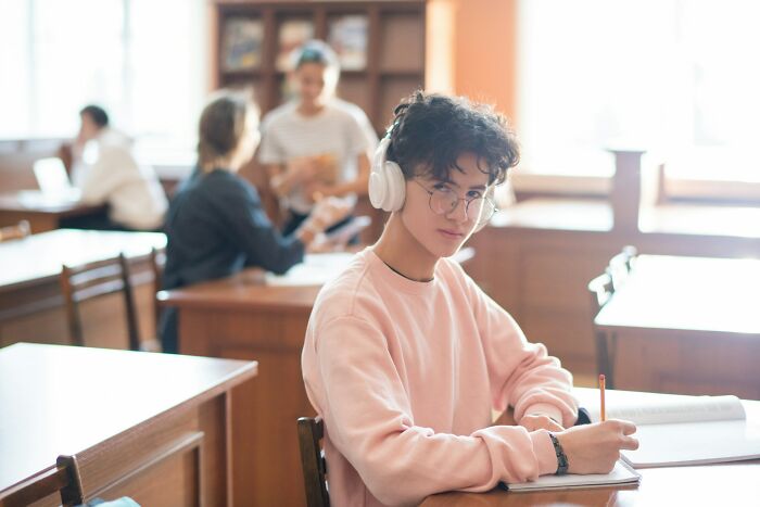 Young person wearing headphones and glasses, writing in notebook in a classroom setting reflecting on backstabbing regrets.