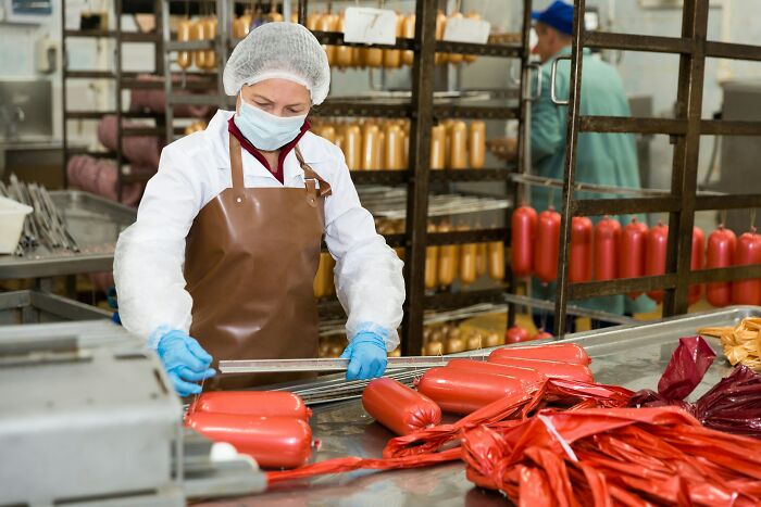 Food industry employee in protective gear handling processed sausages, revealing dark, dirty truths about industries.