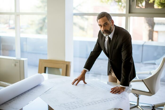 Middle-aged man in a suit looking serious while leaning on architectural plans, reflecting on dark secrets revealed.