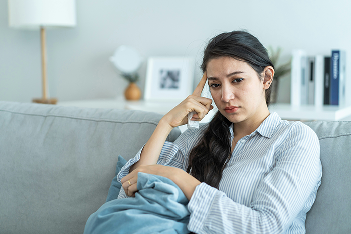 Mother sitting on a couch looking thoughtful and concerned, holding a blanket in a calm home setting. Mother sitting on a couch looking thoughtful and concerned, holding a blanket in a calm home setting.