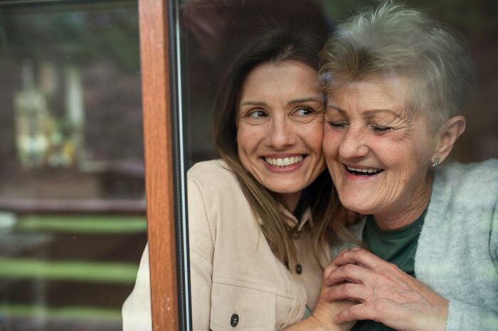 Two women smiling and embracing by a window, representing incredible minds who proved you don’t need a degree to be a genius.