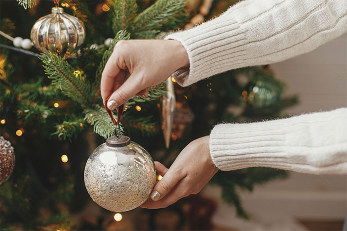 Hands of a woman in a white sweater hanging a silver Christmas ornament on a decorated tree during the holiday season. Hands of a woman in a white sweater hanging a silver Christmas ornament on a decorated tree during the holiday season.