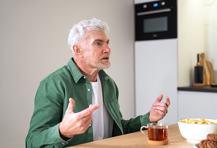 Elderly man in green shirt at kitchen table talking while preparing to wake up granddaughter and make food.