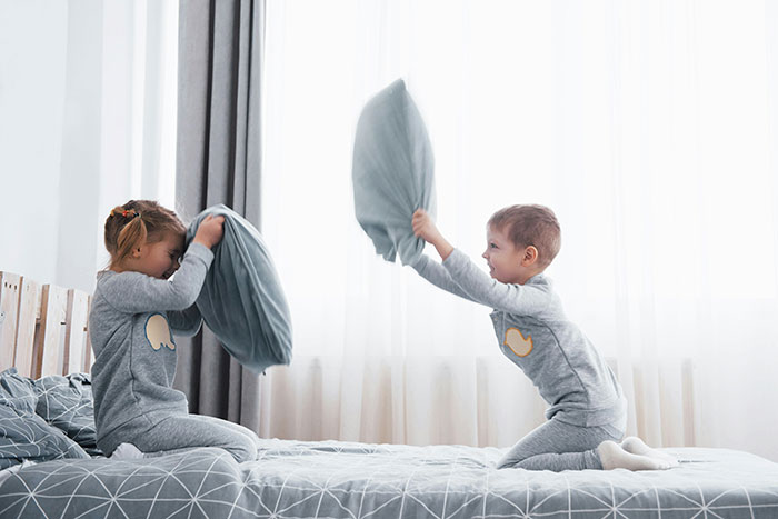 Two young siblings in gray pajamas having a playful pillow fight on a bed in a bright bedroom.