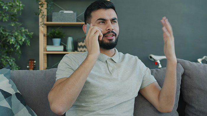 Man with beard sitting on couch, talking on phone with frustrated expression, illustrating gay bro treats sis like dirt. Man with beard sitting on couch, talking on phone with frustrated expression, illustrating gay bro treats sis like dirt.