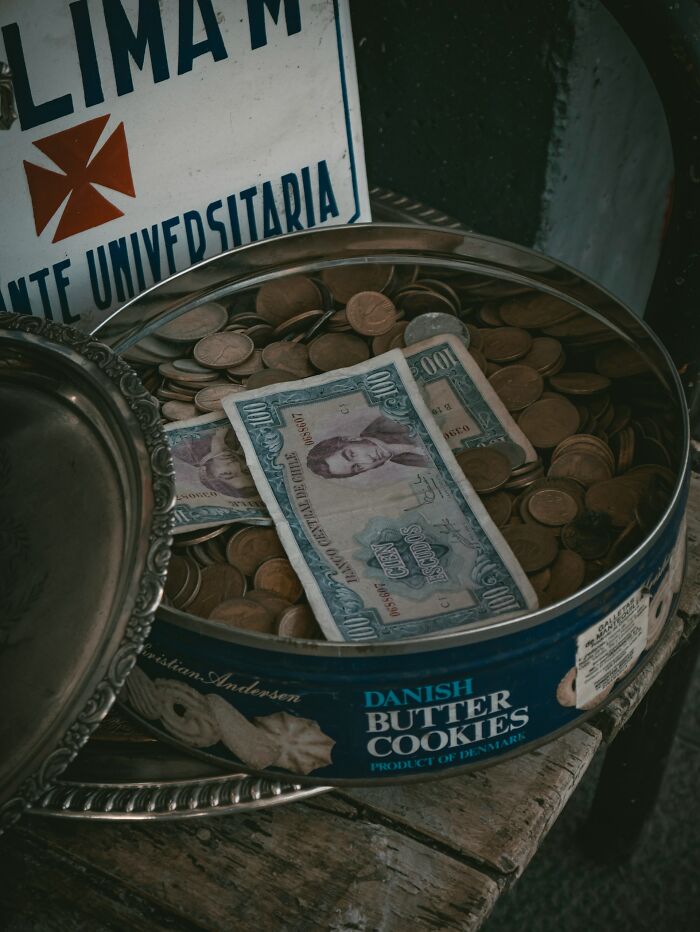 Old currency notes and coins in a vintage Danish butter cookies tin, symbolizing disturbing facts about money people learned.